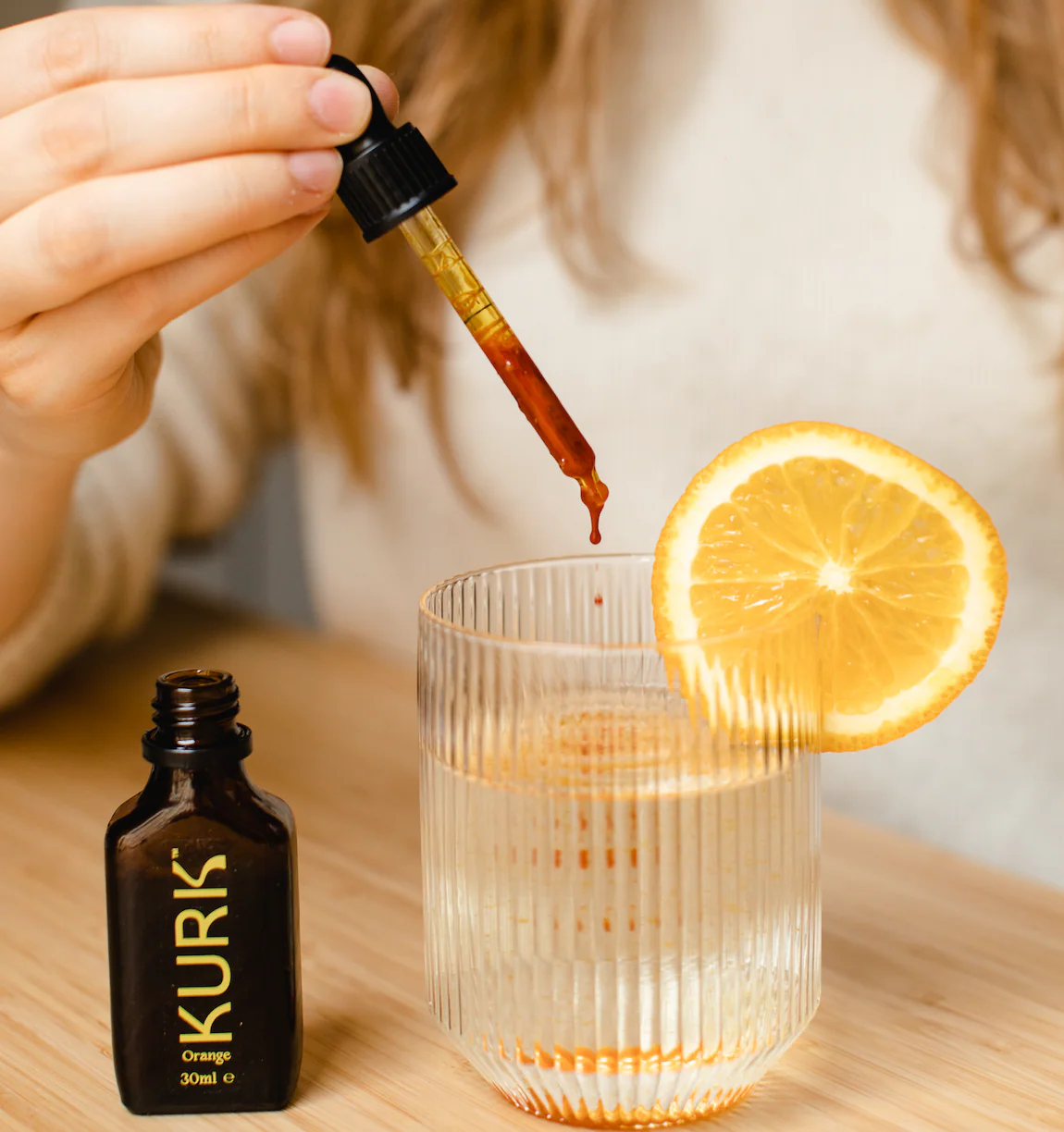 Person adding orange syrup from a dropper bottle into a glass with an orange slice on a wooden table.
